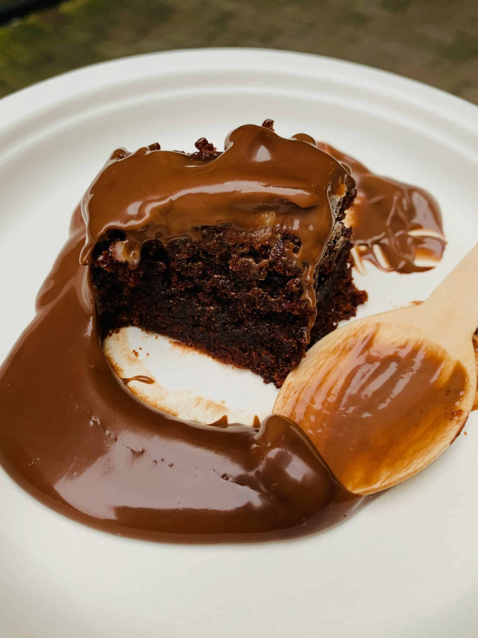 A slice of rich chocolate cake partially covered in glossy chocolate sauce sits on a white compostable paper plate. A wooden spoon with chocolate sauce rests beside it, suggesting the dessert is ready to be enjoyed.