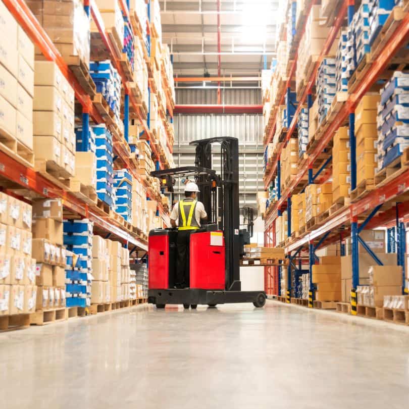 Distributor Warehouse A worker in a high-visibility vest operates a red forklift in a large warehouse aisle, surrounded by tall shelves filled with stacked cardboard boxes and packages.
