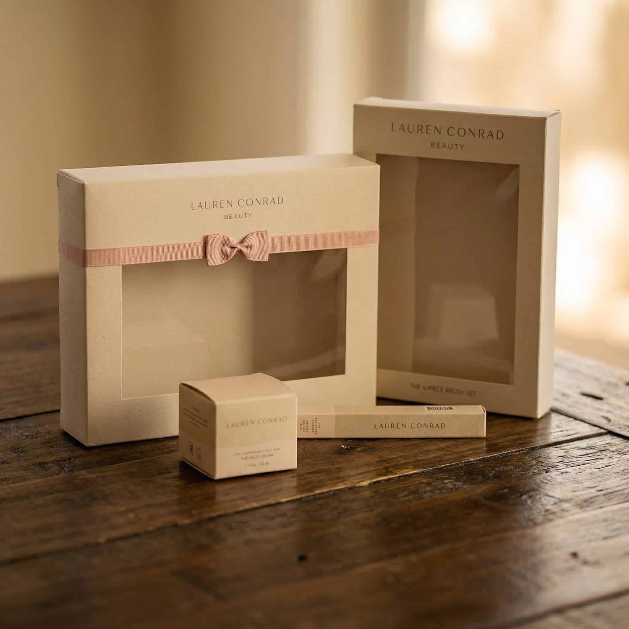 Four beige Lauren Conrad Beauty product boxes, including a brush set and makeup items, are arranged on a rustic wooden surface. One box is decorated with a pink ribbon bow.