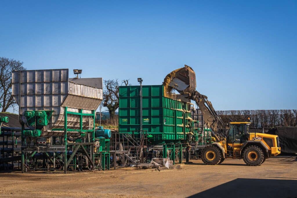 A yellow JCB loader lifts and dumps material into a large green industrial machine at a composting facility